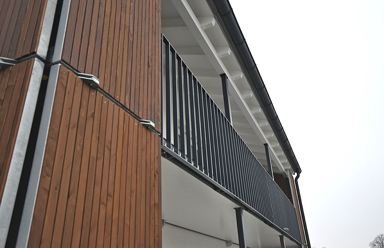 Blick entlang der oberen Etage eines modernen Wohngebäudes mit vertikaler Holzverkleidung, überdachtem Balkon und dunklem Geländer.