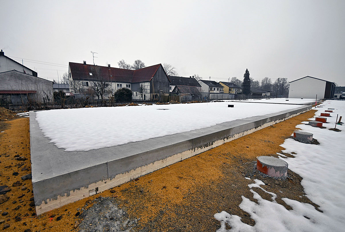 Eine rechteckige Bodenplatte für ein Wohngebäude liegt auf einem verschneiten Baugrundstück. Im Hintergrund sind benachbarte Wohnhäuser zu sehen.