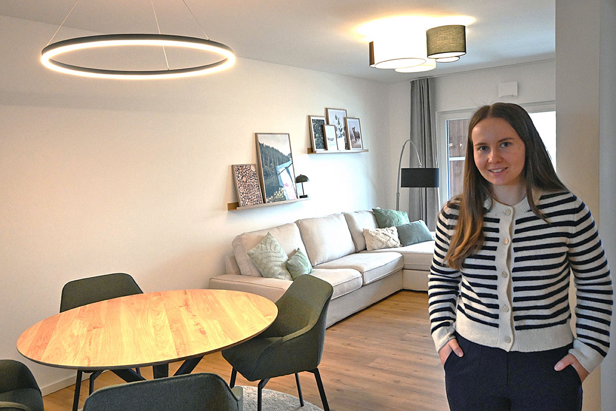 Eine Person steht in einem hellen Wohn- und Essbereich mit rundem Esstisch, Ecksofa und moderner Einrichtung. An der Wand hängen Bilder auf schmalen Regalen.