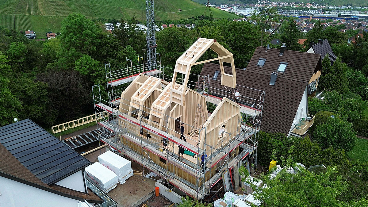 Holzhaus im Bau: Vorgefertigte Wand- und Dachelemente werden mit einem Kran auf einem eingerüsteten Gebäude montiert.