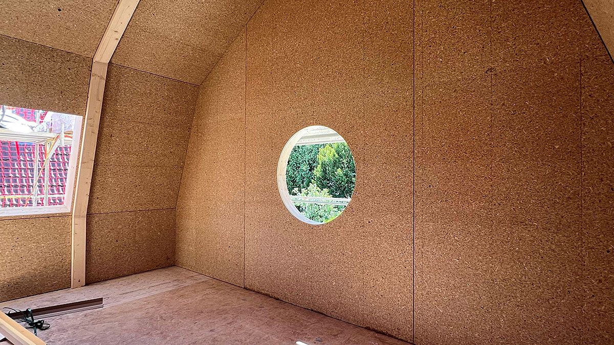 Rohbau eines Dachgeschossraums mit sichtbaren Holzwerkstoffplatten und rundem Fenster in der Giebelwand mit Blick ins Grüne.