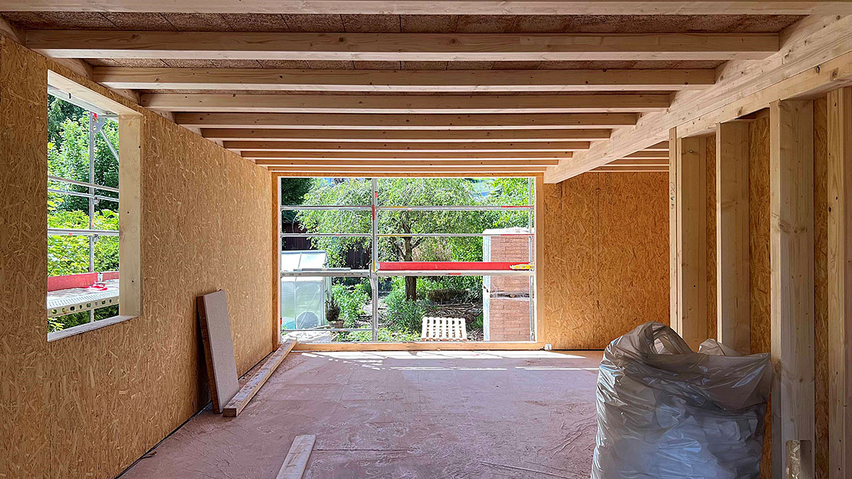 Innenraum eines Holzhauses im Rohbau mit sichtbarer Balkendecke und großer Fensteröffnung zum Garten.