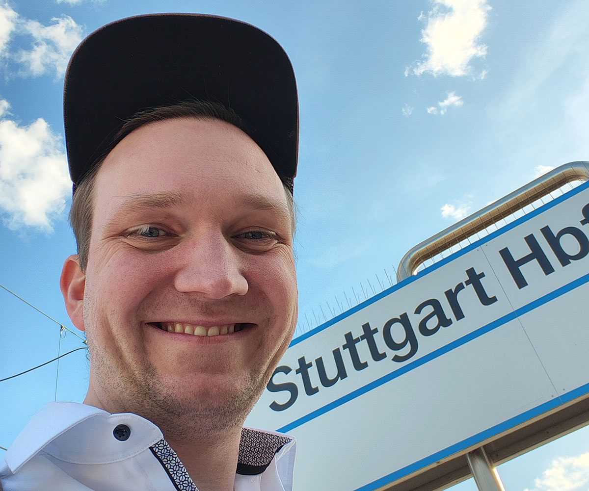 Lächelnde Person mit schwarzer Kappe macht ein Selfie vor einem Bahnhofsschild mit der Aufschrift „Stuttgart Hbf“ unter blauem Himmel.