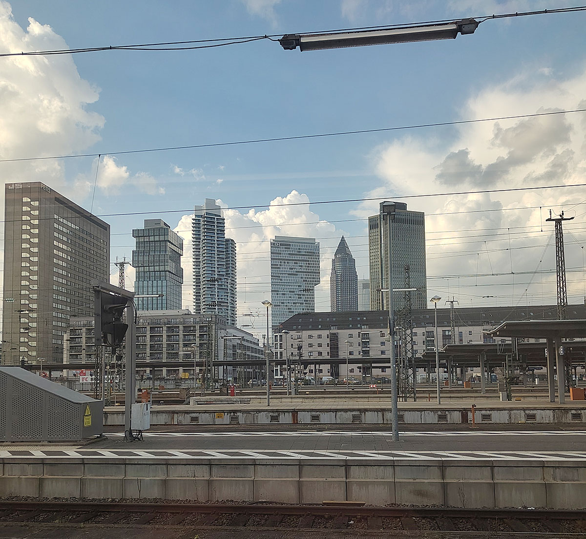 Blick über mehrere Bahngleise und Bahnsteige auf die Hochhäuser der Frankfurter Skyline unter einem teilweise bewölkten Himmel.