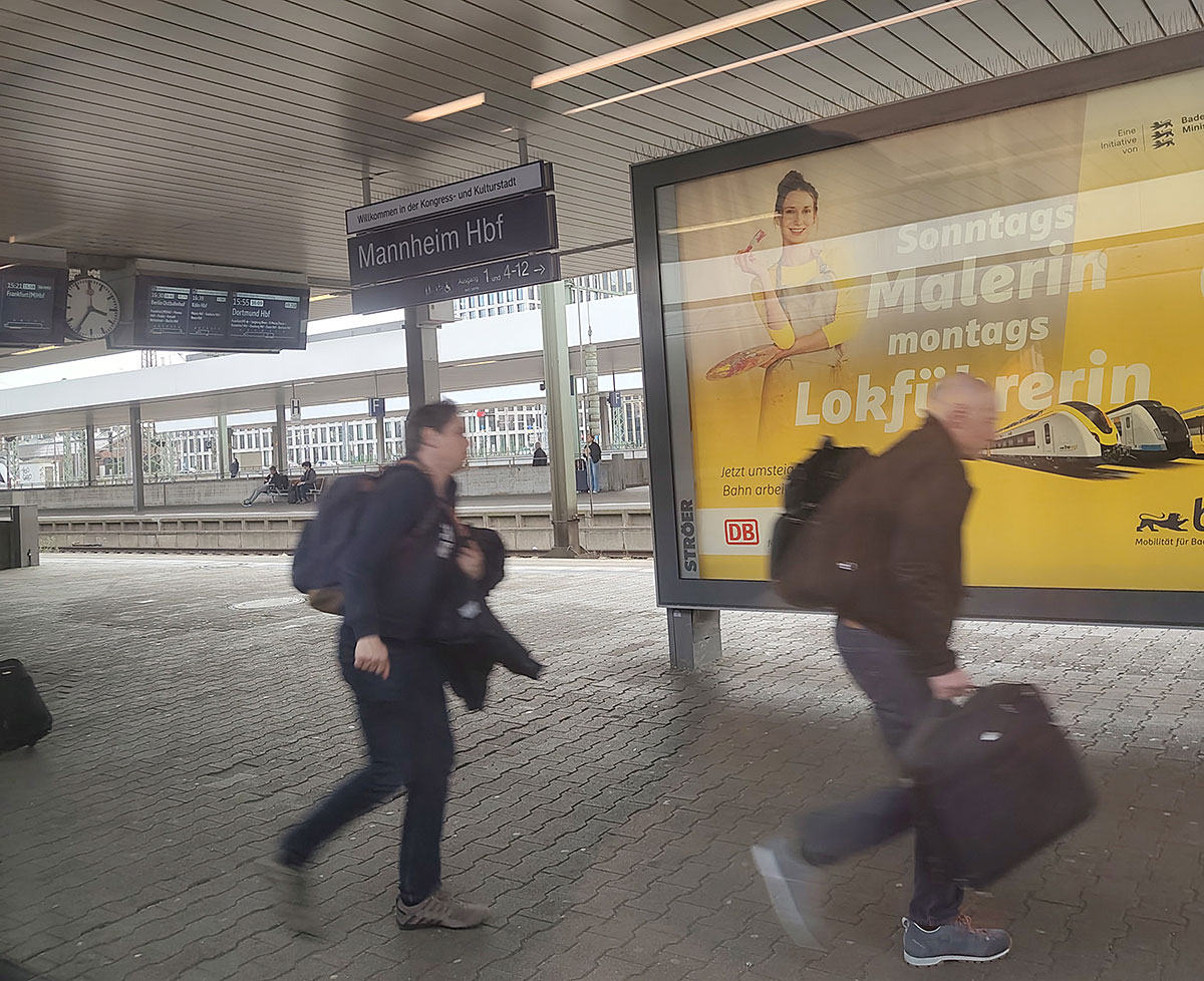 Bahnsteig am Bahnhof Mannheim Hbf mit vorbeigehenden Reisenden, Anzeigetafeln und einem großen Werbeplakat der Deutschen Bahn.