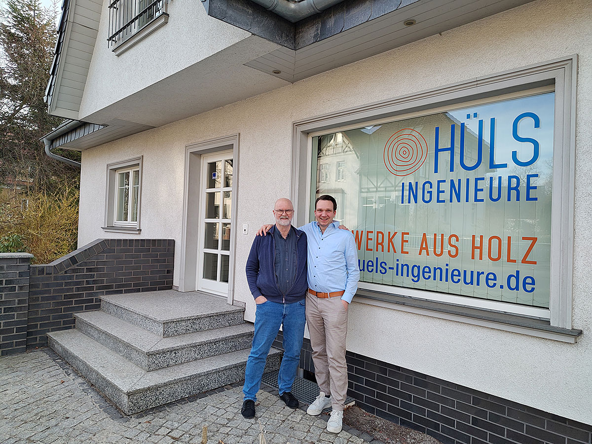 Zwei Personen stehen vor einem Bürogebäude neben einem Fenster mit der Aufschrift „Hüls Ingenieure – Werke aus Holz“.