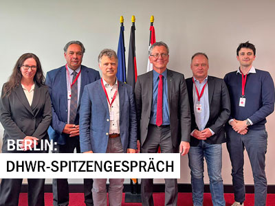 Sechs Personen stehen vor mehreren Flaggen für ein Gruppenfoto. Im Bild eingeblendet ist der Schriftzug ‚Berlin: DHWR-Spitzengespräch‘.