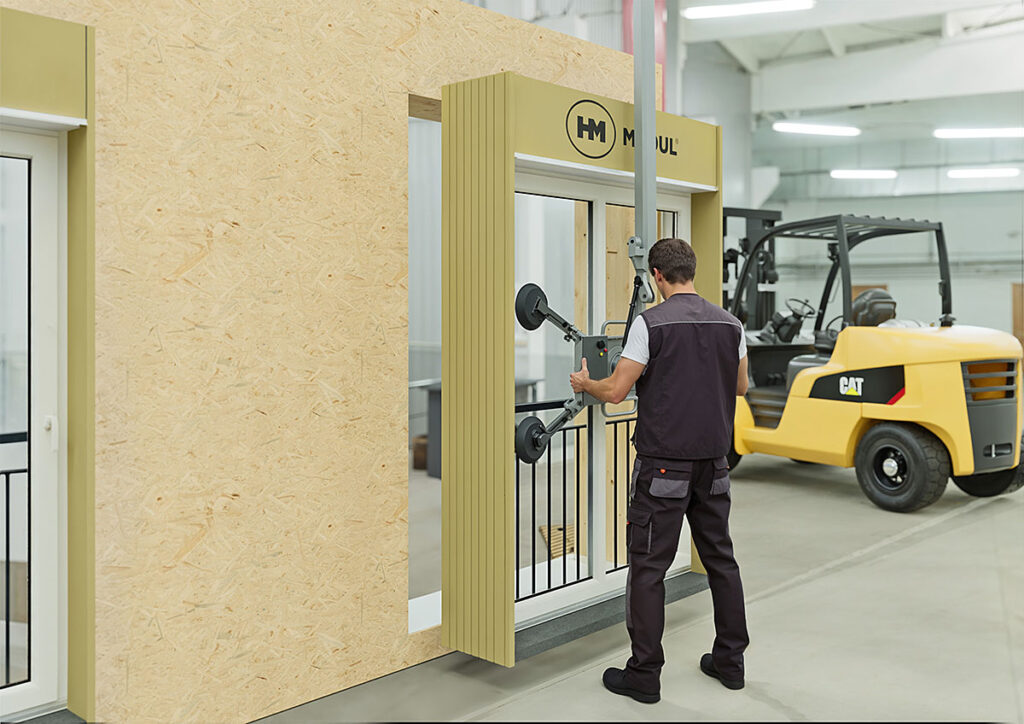 Fenster wird mithilfe eines Vakuumhebers in eine vorbereitete Wandöffnung eingesetzt; eine Person richtet das Element aus, im Hintergrund ein Gabelstapler.