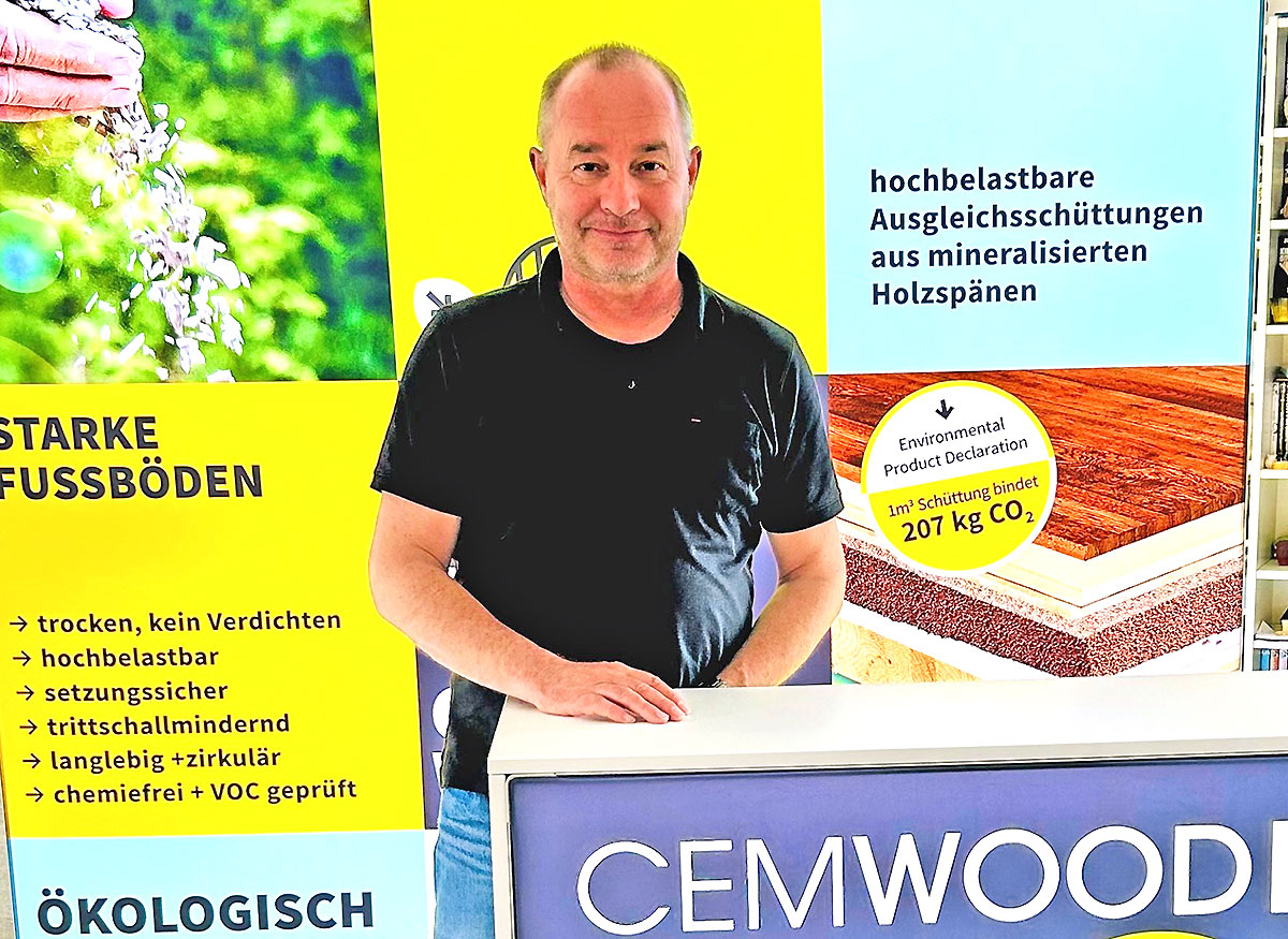 Person steht hinter einem Messestand von CEMWOOD vor großformatigen Werbetafeln zu ökologischen Holzspan-Schüttungen für Fußböden. Im Hintergrund werden Eigenschaften wie hohe Belastbarkeit, Trittschallminderung und chemiefreie Verarbeitung hervorgehoben.