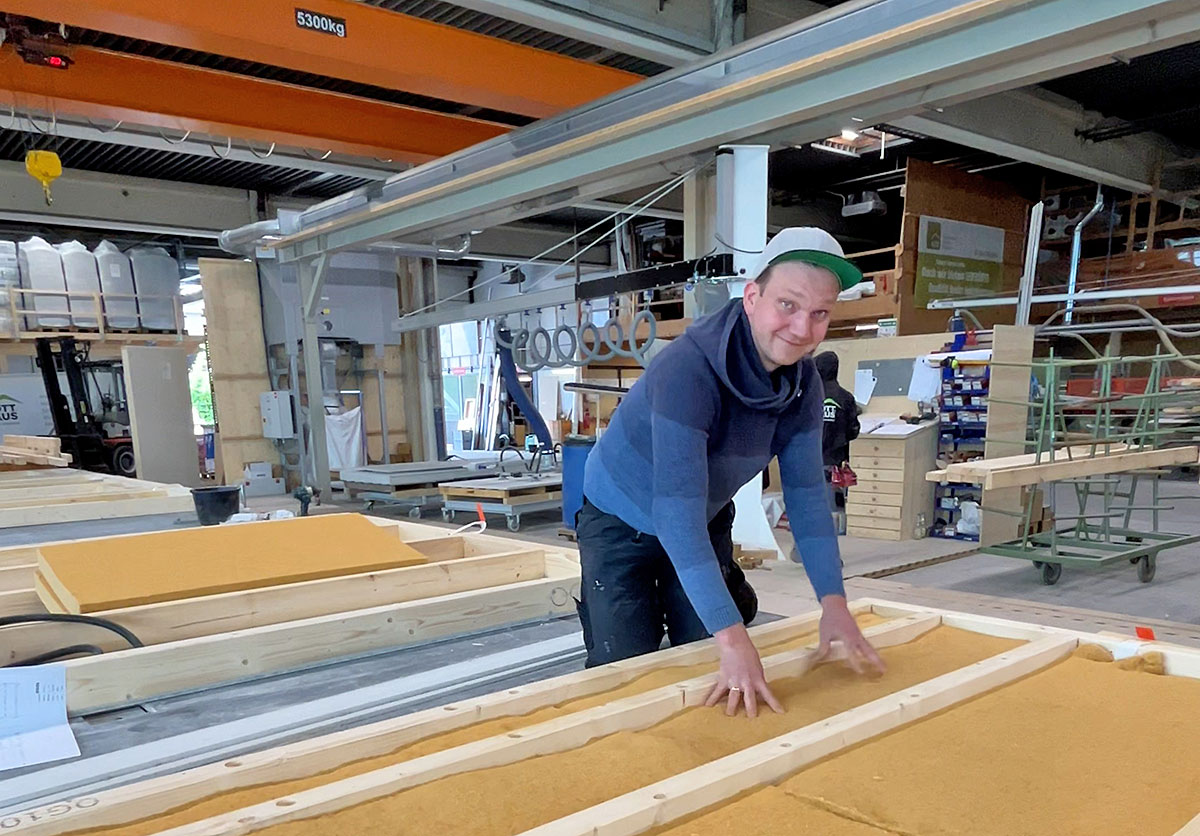 Person mit Kappe arbeitet in einer Holzfertigungshalle an einem offenen Holzrahmen mit Dämmmaterial; im Hintergrund sind Maschinen, Werkbänke und weitere Bauelemente zu sehen.