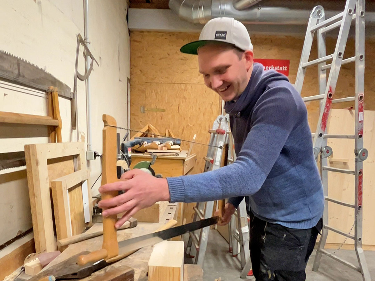 Person mit Kappe arbeitet in einer Werkstatt mit einer Handsäge an einem Holzbauteil; im Hintergrund stehen Leitern, Werkzeuge und weitere Holzelemente.