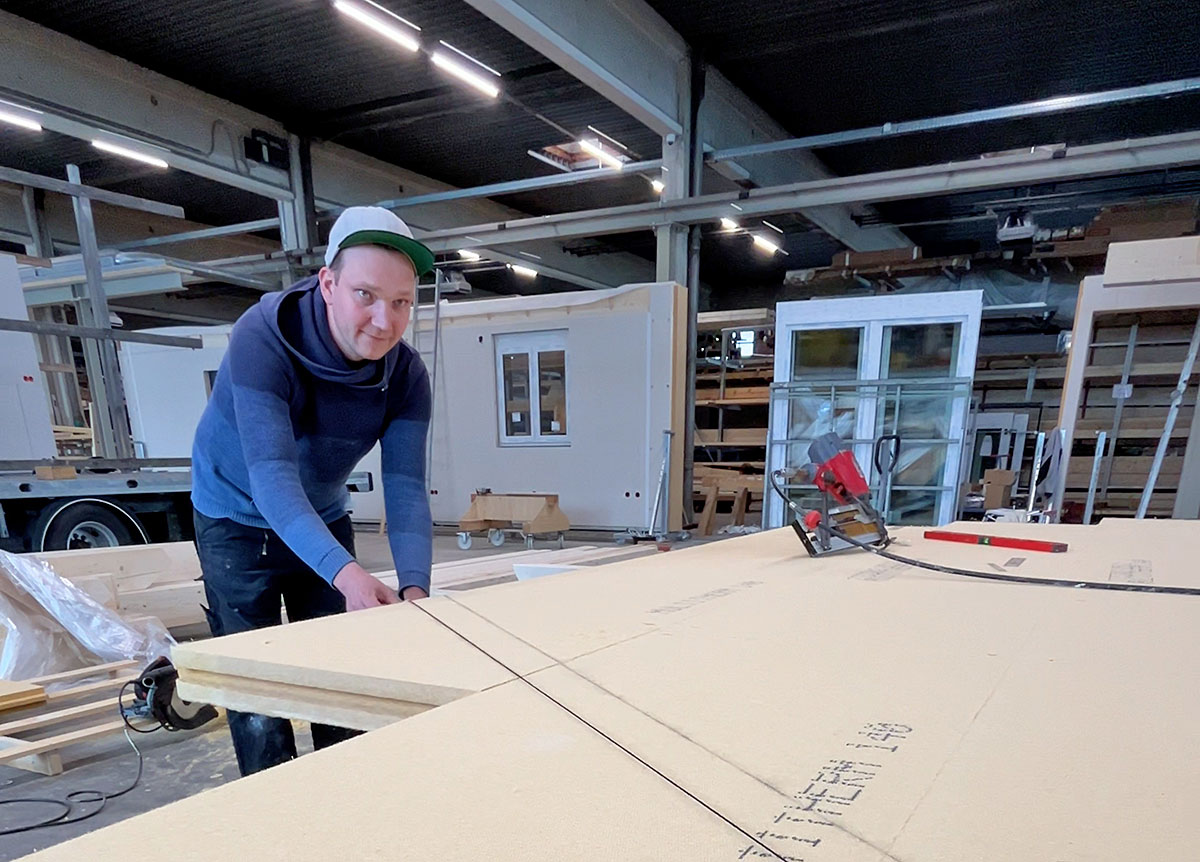 Person mit Kappe arbeitet in einer Fertigungshalle an großen Holzplatten; im Hintergrund sind vorgefertigte Wandelemente, Fensterrahmen und Werkzeuge zu sehen.