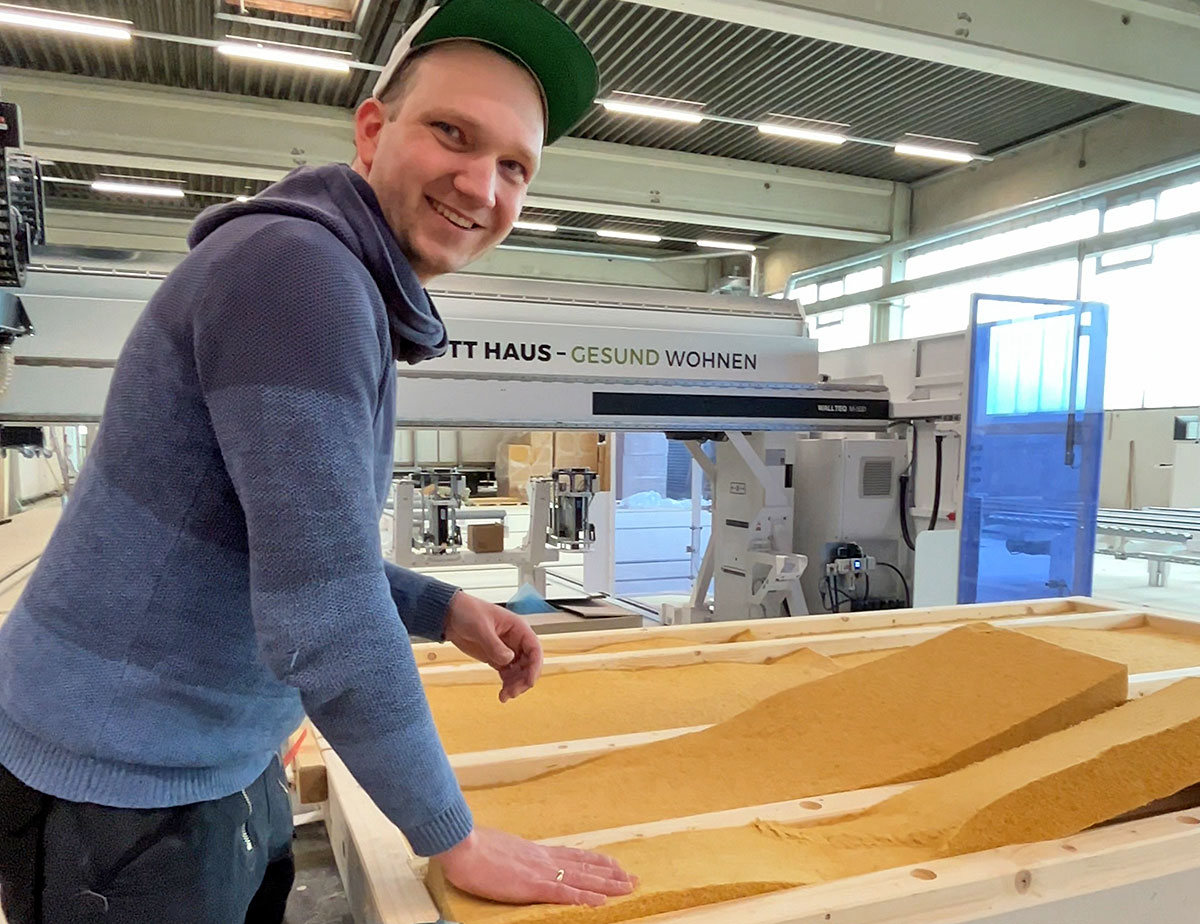 Person mit grüner Kappe lehnt an einer Holzrahmenkonstruktion in einer Produktionshalle; im Hintergrund ist eine Fertigungsanlage von „OTT HAUS – Gesund wohnen“ zu sehen. Eingeblendeter Text: „Reisetagebuch – Auf zu OTT Haus“.