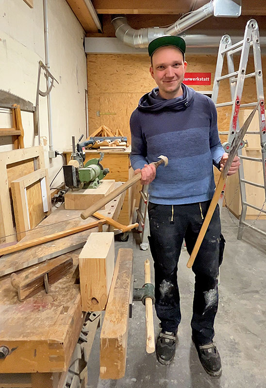 Person mit grüner Kappe steht in einer Werkstatt zwischen Werkbank und Leiter und hält zwei Handwerkzeuge aus Holz und Metall in den Händen.