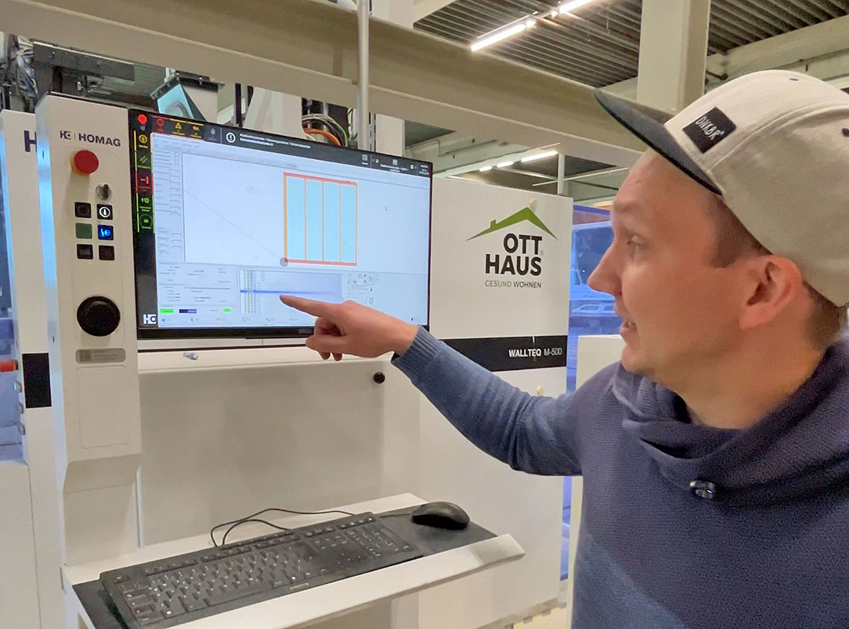 Person mit Kappe zeigt in einer Produktionshalle auf einen Bildschirm mit digitalem Bauplan an einer Maschinensteuerung von „OTT HAUS – Gesund wohnen“.