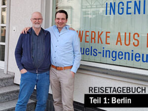 Zwei Personen stehen nebeneinander vor einem Schaufenster mit der Aufschrift eines Ingenieurbüros. Unten im Bild steht der Titel „Reisetagebuch – Teil 1: Berlin“.