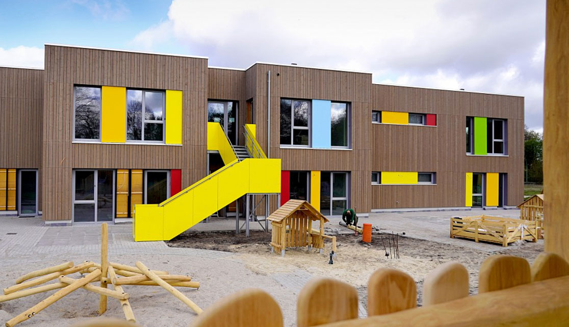 Zweigeschossiges Gebäude mit Holzfassade und farbigen Fenster- und Fassadenelementen. Im Innenhof befinden sich Spielgeräte und Sandflächen, die auf eine Kindertagesstätte oder Bildungseinrichtung hinweisen.