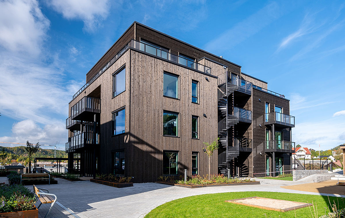 Mehrgeschossiges Wohngebäude mit dunkler Holzfassade, großen Fensterflächen und Balkonen. Eine außenliegende Treppe verbindet die Etagen, vor dem Gebäude befinden sich Grünflächen und Aufenthaltsbereiche.