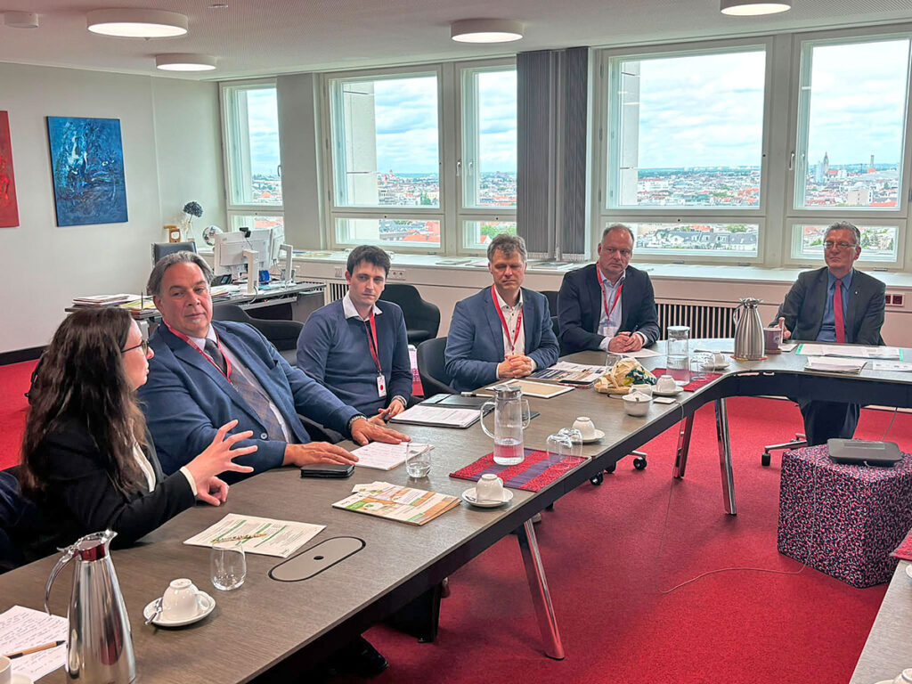 Mehrere Personen sitzen in einem Besprechungsraum an einem U-förmig angeordneten Konferenztisch und diskutieren miteinander. Im Hintergrund sind große Fenster mit Blick über die Stadt zu sehen.